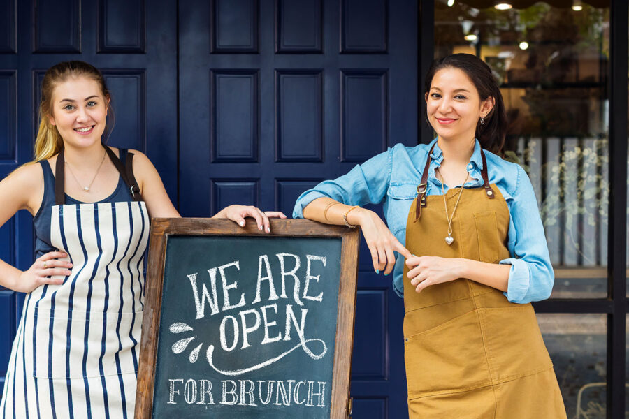 Two women standing in front of a blue doorway to their business with elbows resting on a sandwich board that says WE ARE OPEN