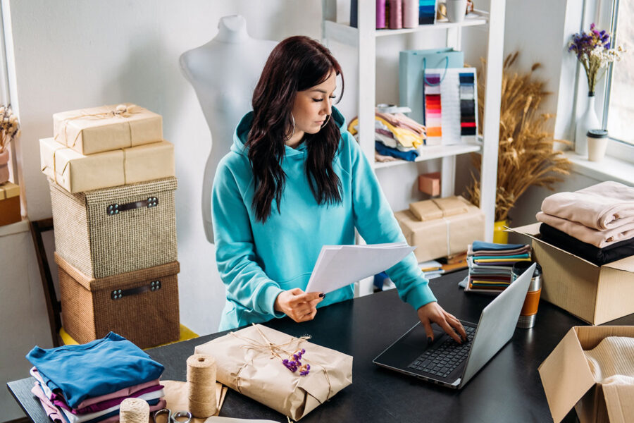 Woman surrounded by boxes and cloth material holding papers while typing on a laptop