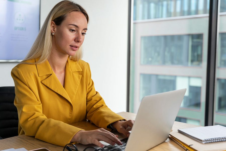 Blonde business woman in gold suit typing on a laptop with a window in the background